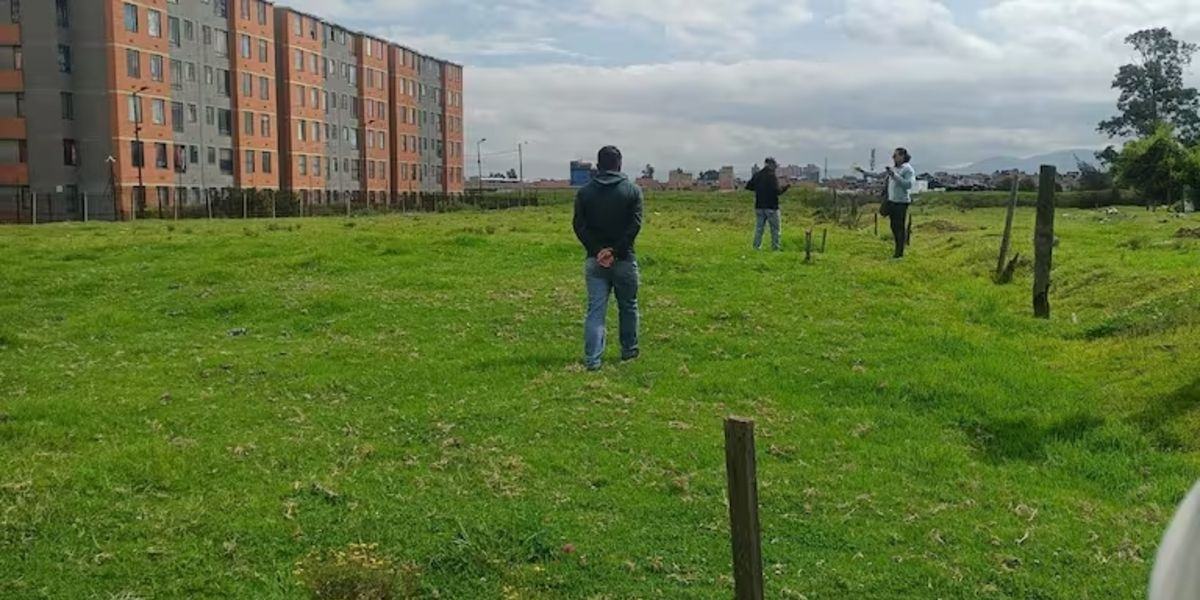 Tres personas permanecen sobre un terreno con césped junto a edificios de apartamentos y una cerca bajo un cielo nublado.