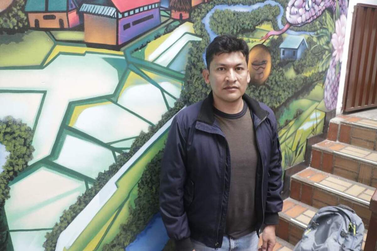 Persona de pie frente a un mural colorido con paisajes y una serpiente. A la derecha hay una escalera y una mochila en el suelo.