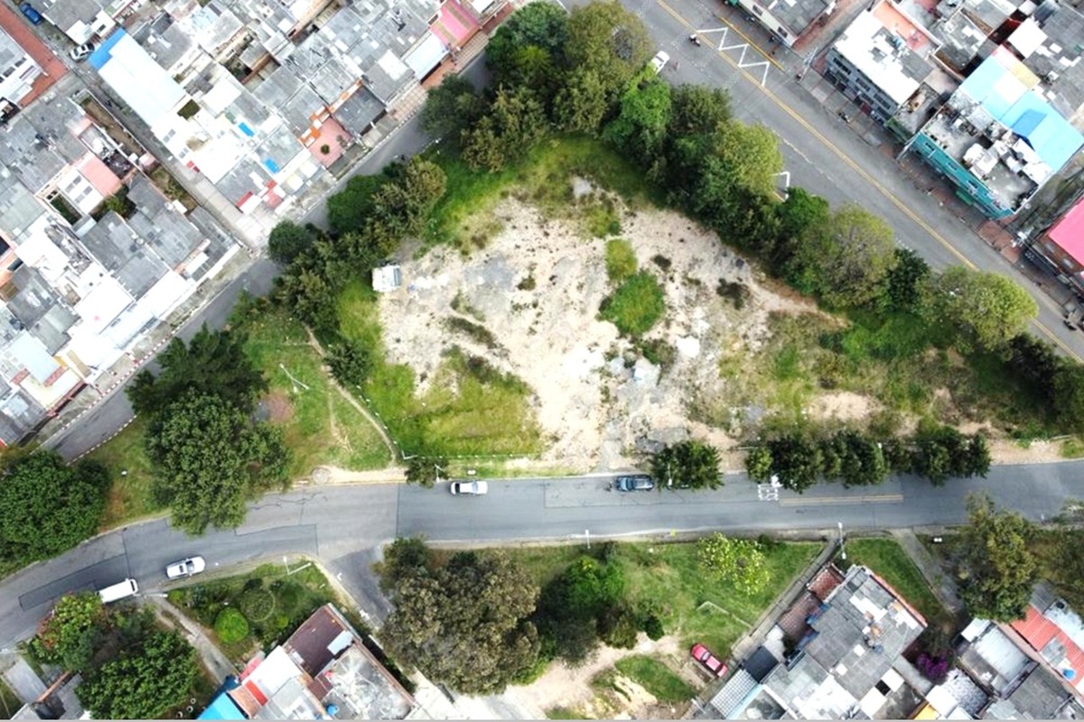 Vista aérea de un lote baldío con pasto y tierra, rodeado por árboles. Calles con autos lo delimitan y hay casas densamente construidas en los bordes.		