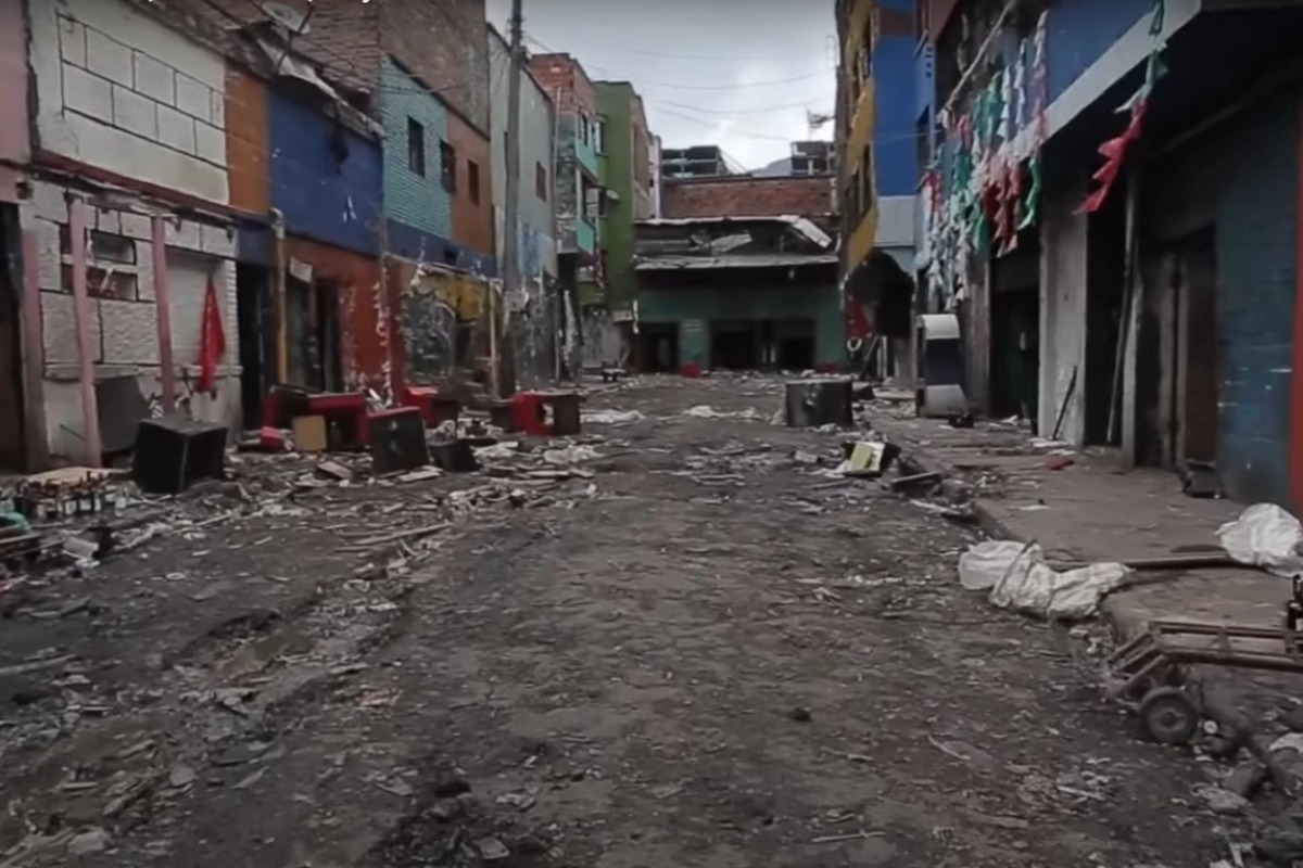 Calle estrecha y vacía entre edificios de varios pisos y colores, con fachadas deterioradas, grafitis y banderines; suelo cubierto de escombros, basura, cajas y objetos tirados.		