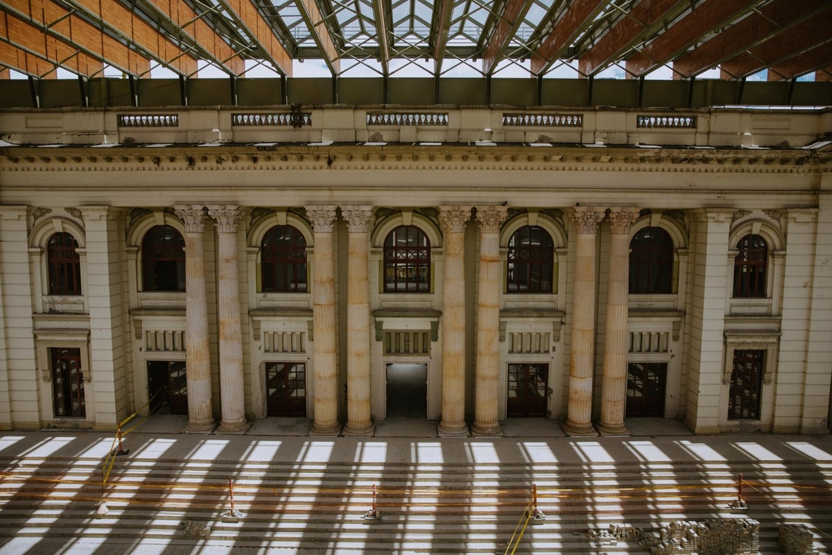 Edificio clásico interior con altos pilares y ventanas arqueadas suelo con amplias escaleras iluminadas por sombras de estructura metálica superior zona en reparación con cintas y escombros.