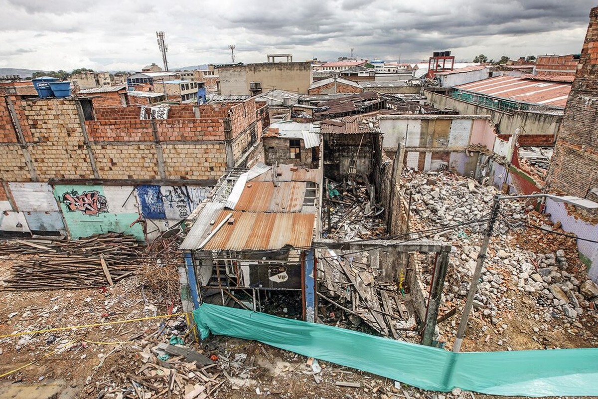 Vista aérea de zona urbana degradada: casas de ladrillo y techos de metal, edificio derrumbado con escombros y maderas, grafitis en muros y cinta verde delimitando el área.		