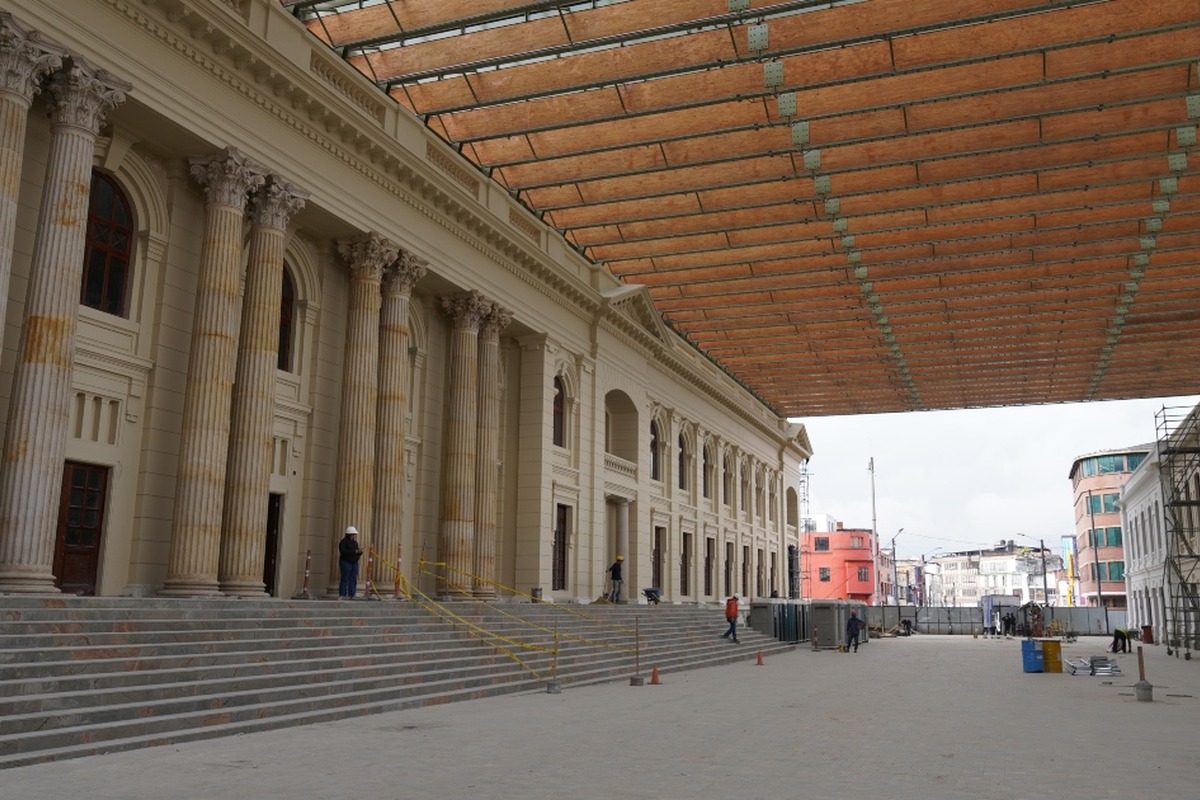 Edificio clásico con columnas y escalinata en restauración varios obreros con cascos trabajan. Gran techo provisional cubre la calle al fondo se ven más edificios urbanos.