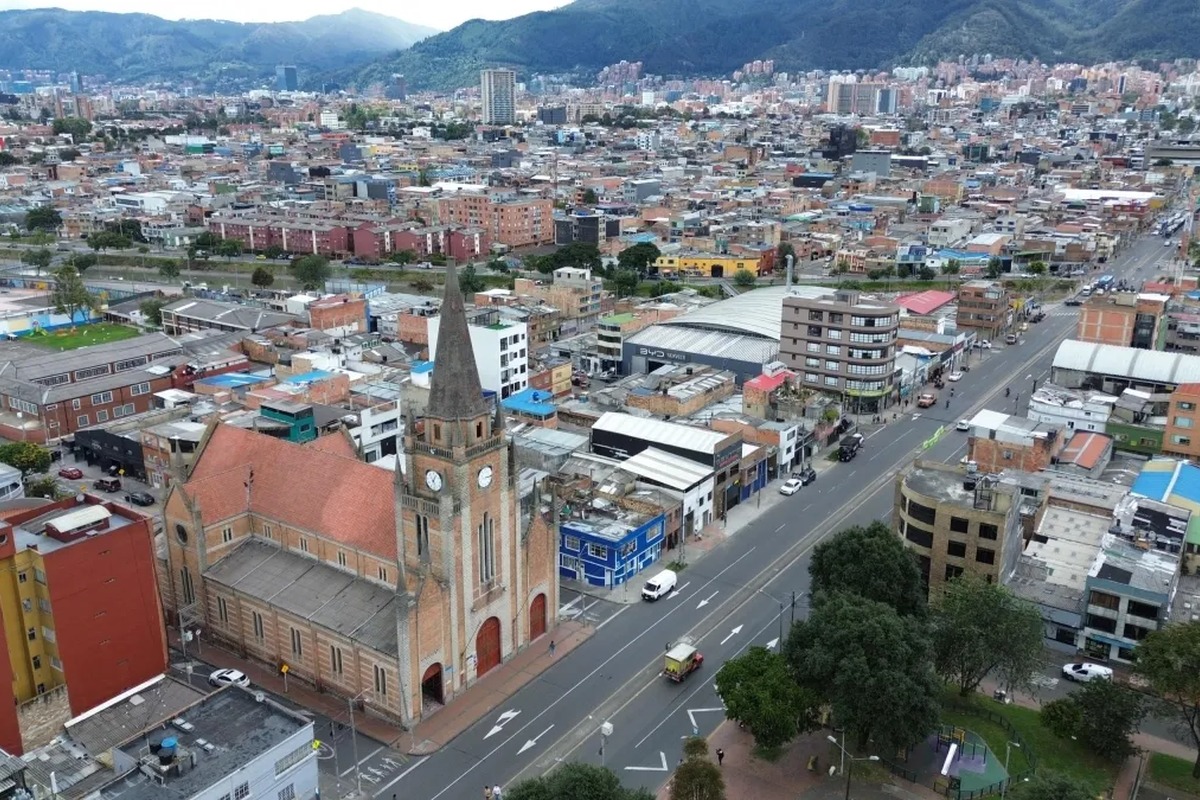 Vista aérea de ciudad con muchos edificios bajos, avenida principal con autos, iglesia grande de ladrillo con torre y reloj, parque con árboles y montañas al fondo.