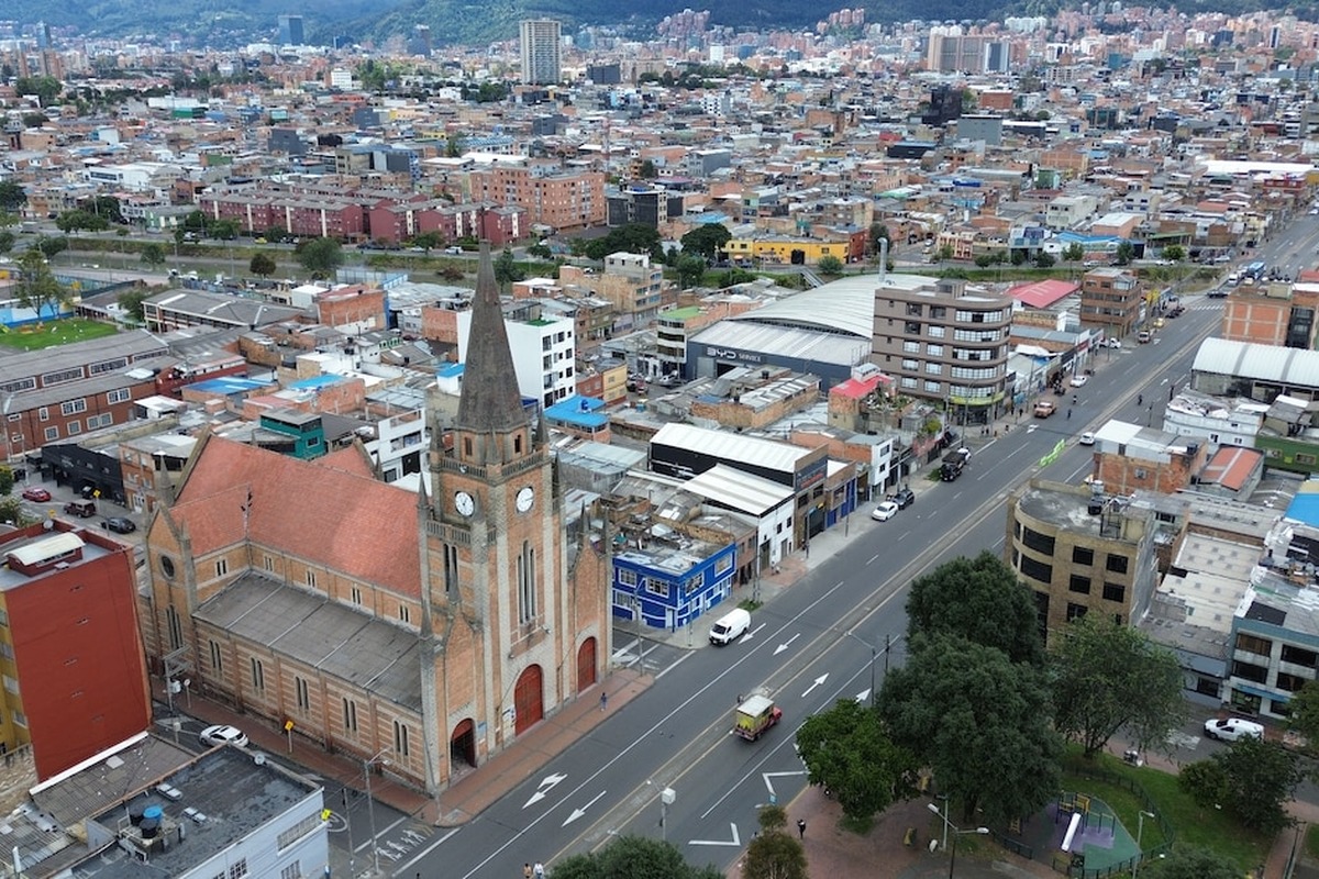 Vista aérea de ciudad densa; destaca iglesia grande de ladrillo con torre y reloj junto a avenida con pocos vehículos, edificios bajos variados y zona verde con árboles y parque infantil.