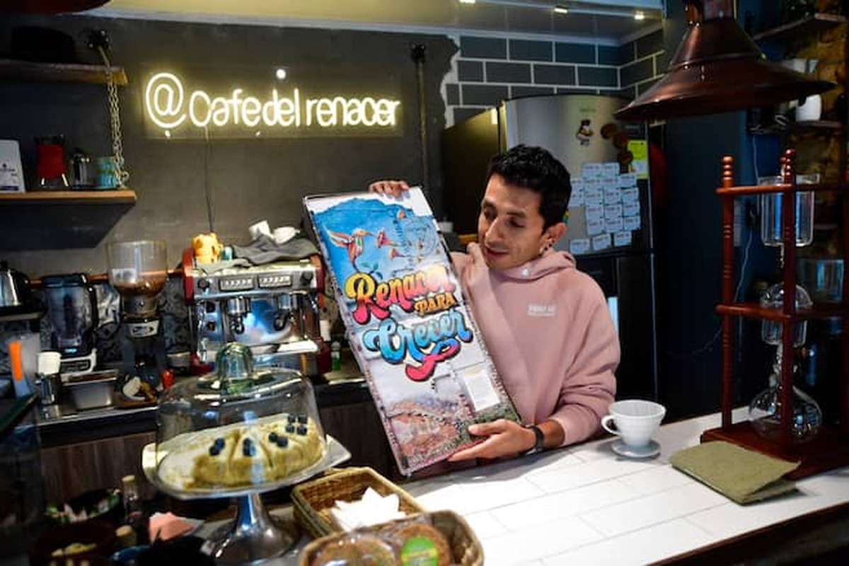 Hombre tras barra de cafetería sostiene un cartel colorido con texto “Renacer para Crecer”. Al fondo máquina de café y letrero “@cafedelrenacer”. Sobre la barra pastel y utensilios.