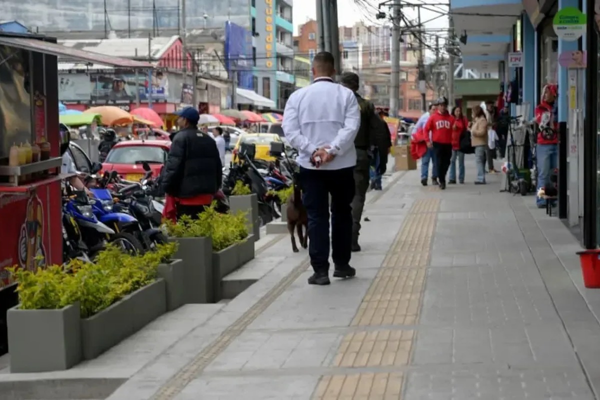Acera urbana con franja táctil amarilla; varias personas caminan, una con perro. A la izquierda hay motos estacionadas, carros, puestos con sombrillas de colores y locales comerciales.