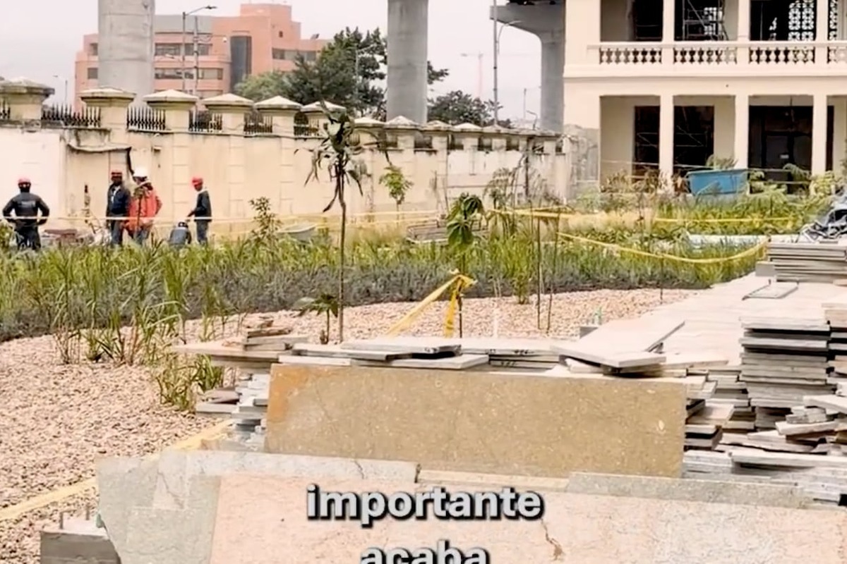 Obra al aire libre con montones de placas de piedra en primer plano, plantas y grava; al fondo varias personas con cascos y edificio en construcción. Texto parcial: “importante acaba”.