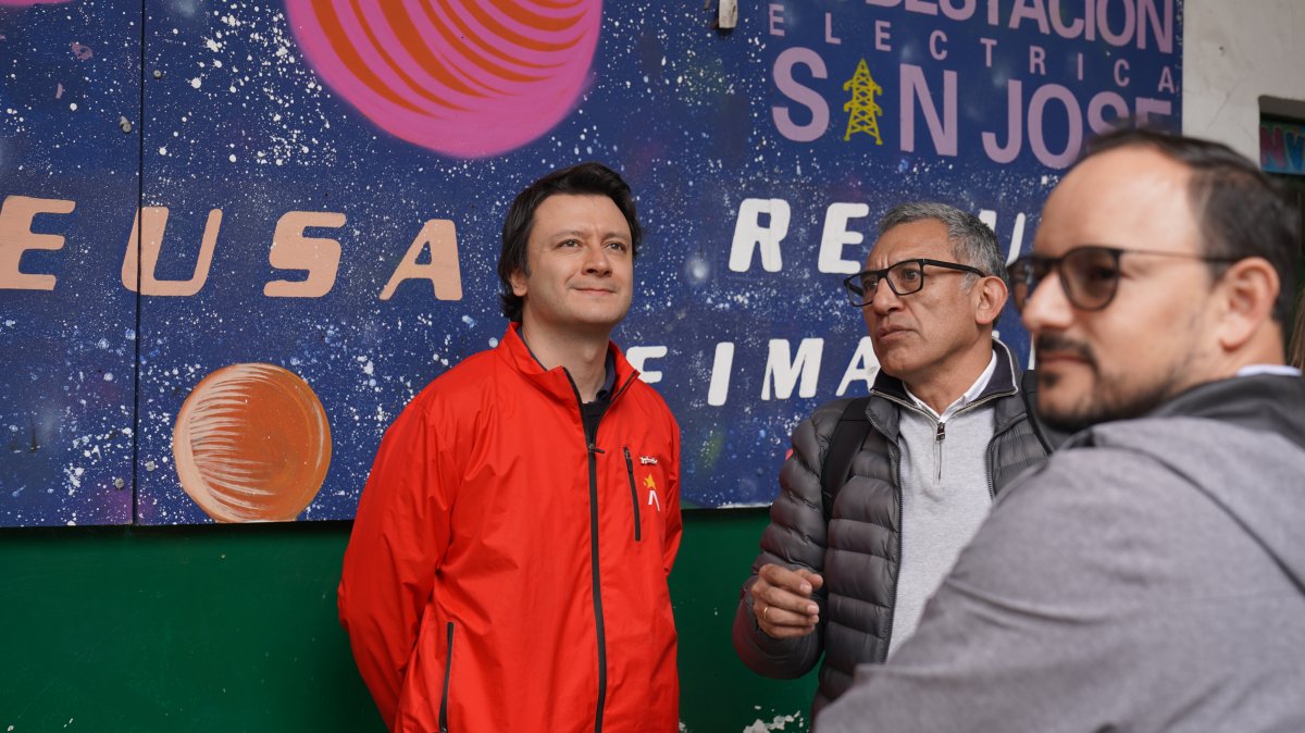 Tres hombres frente a un mural; uno viste chaqueta roja y otro sostiene un micrófono.