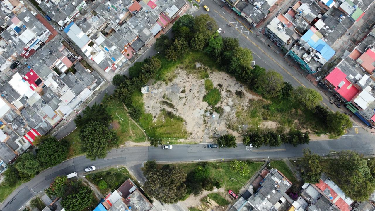 Vista aérea de un lote sin construir, con vegetación, árboles, calles, viviendas y varios vehículos alrededor.