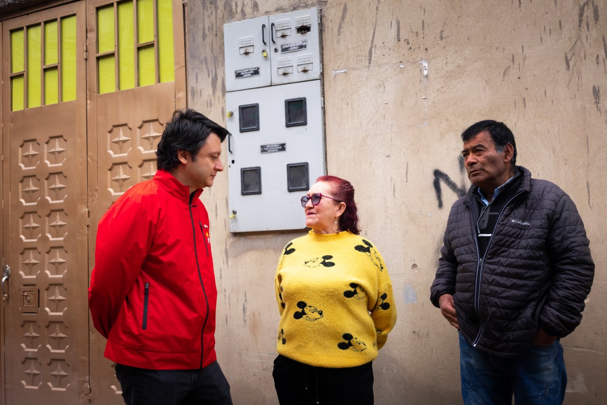 Tres personas conversan frente a una fachada; una viste rojo, otra amarillo y otra una chaqueta negra
