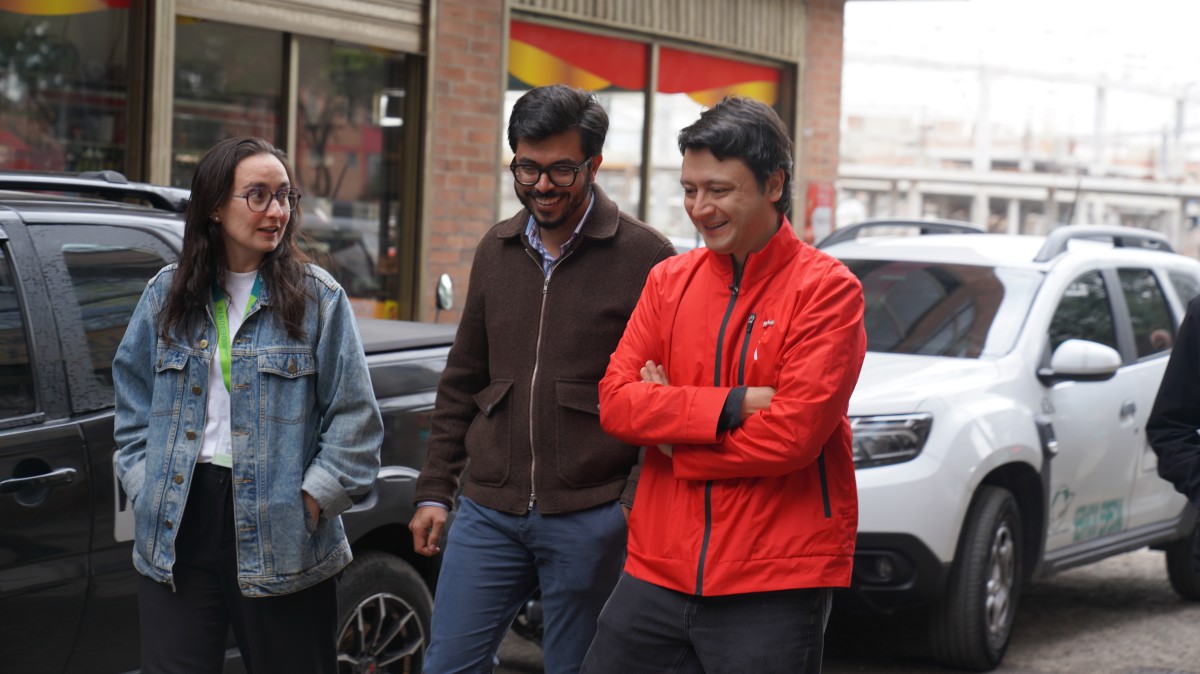 Tres personas caminan junto a vehículos; una lleva chaqueta roja, otra marrón y otra chaqueta de mezclilla.