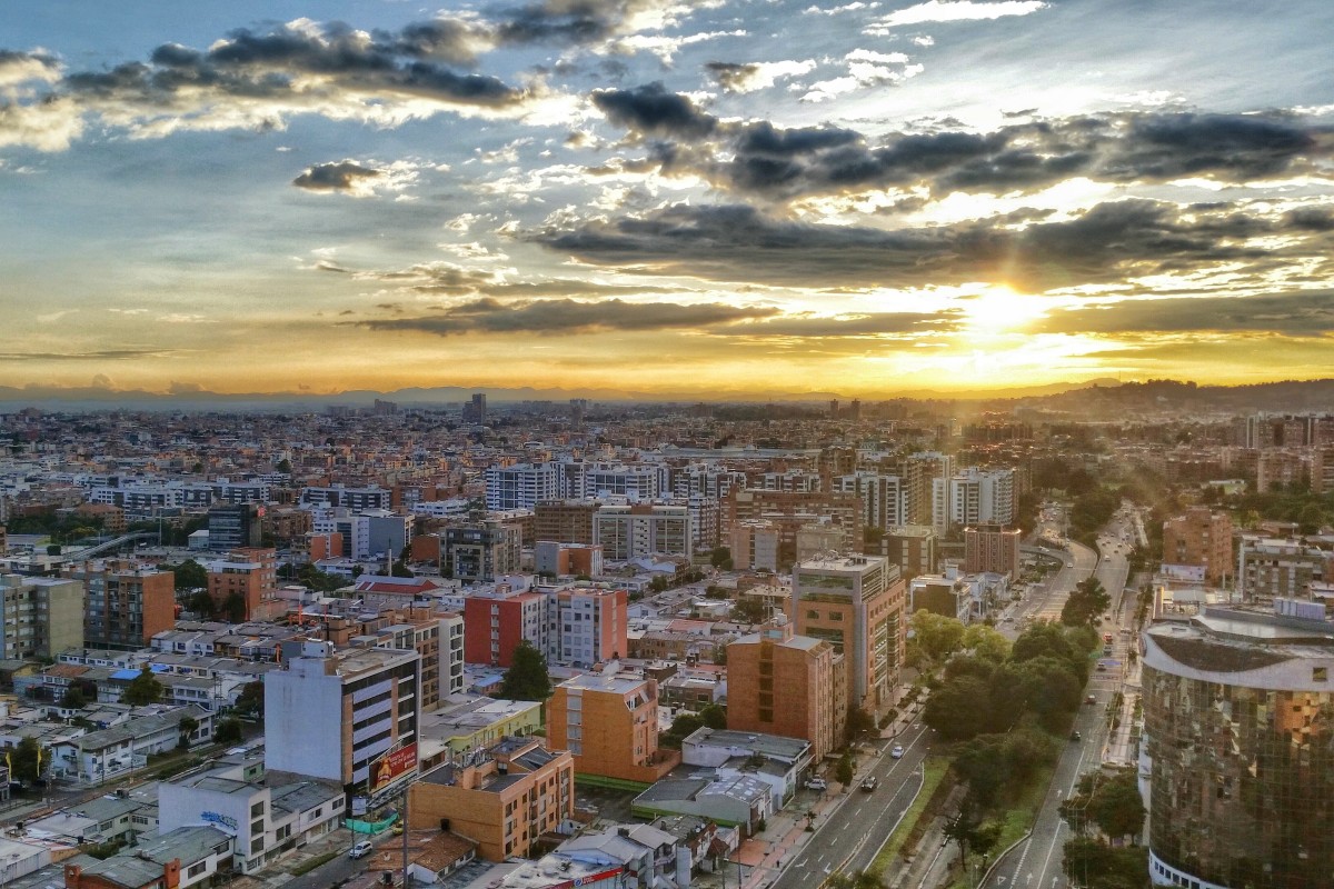 Vista panorámica de una ciudad con edificios, calles y árboles; al fondo, el sol se oculta bajo nubes.