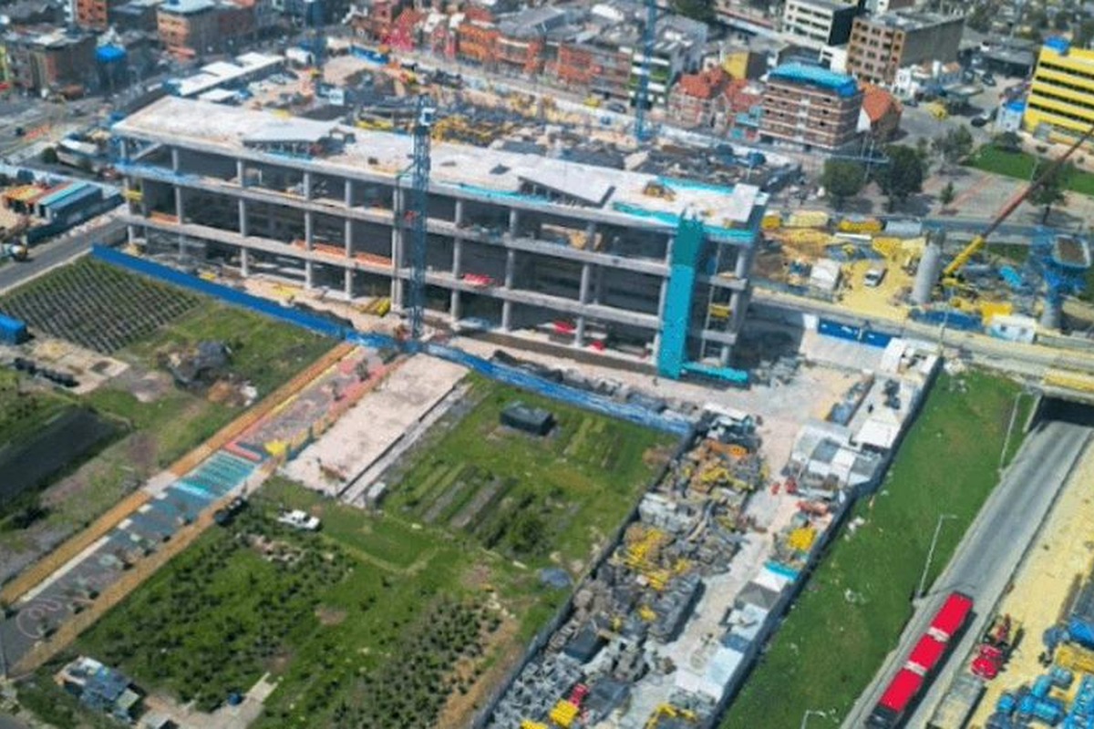 Vista aérea de una gran obra: edificio gris de varios pisos en construcción, grúas, materiales esparcidos, zonas verdes con cultivos y un par de buses rojos en carretera cercana.