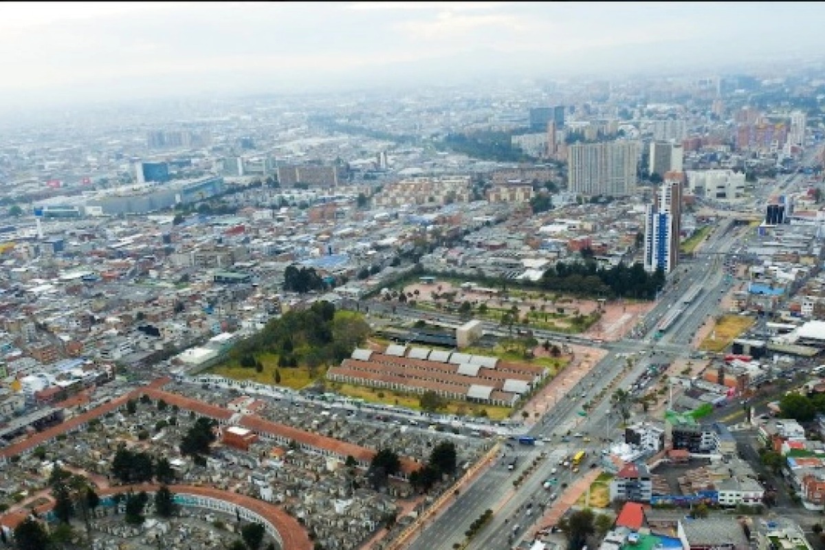 Foto aérea de ciudad grande con muchos edificios bajos y algunos altos, avenidas amplias con vehículos, zonas verdes rectangulares y cielo nublado al fondo.