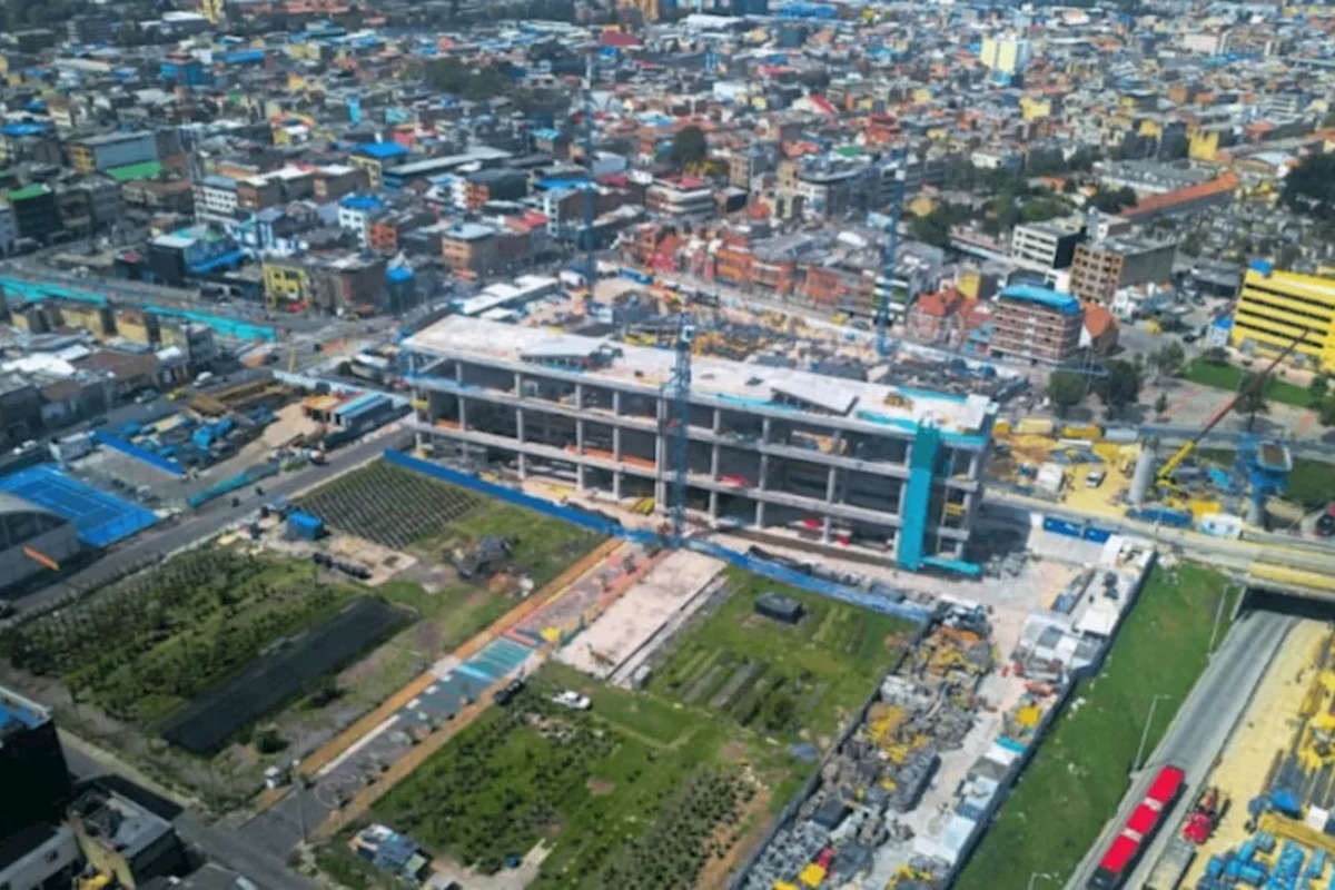 Vista aérea de una ciudad: en el centro, gran edificio en construcción de varios pisos; alrededor hay calles, casas densas, zonas verdes, maquinaria, materiales y una vía con buses rojos.