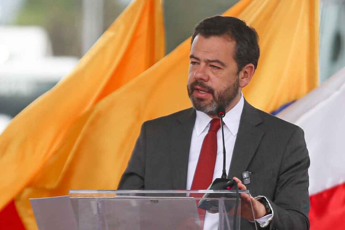 Hombre de traje gris y corbata roja habla en un podio transparente con micrófono detrás hay grandes banderas amarilla y blanca desenfocadas.