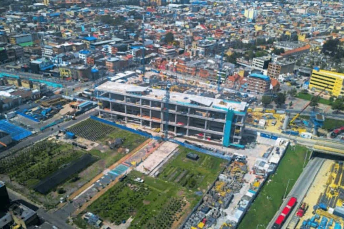 Vista aérea de una ciudad con un gran edificio en construcción en el centro, rodeado de andamios, grúas, material de obra, zonas verdes cultivadas y muchas casas y calles alrededor.