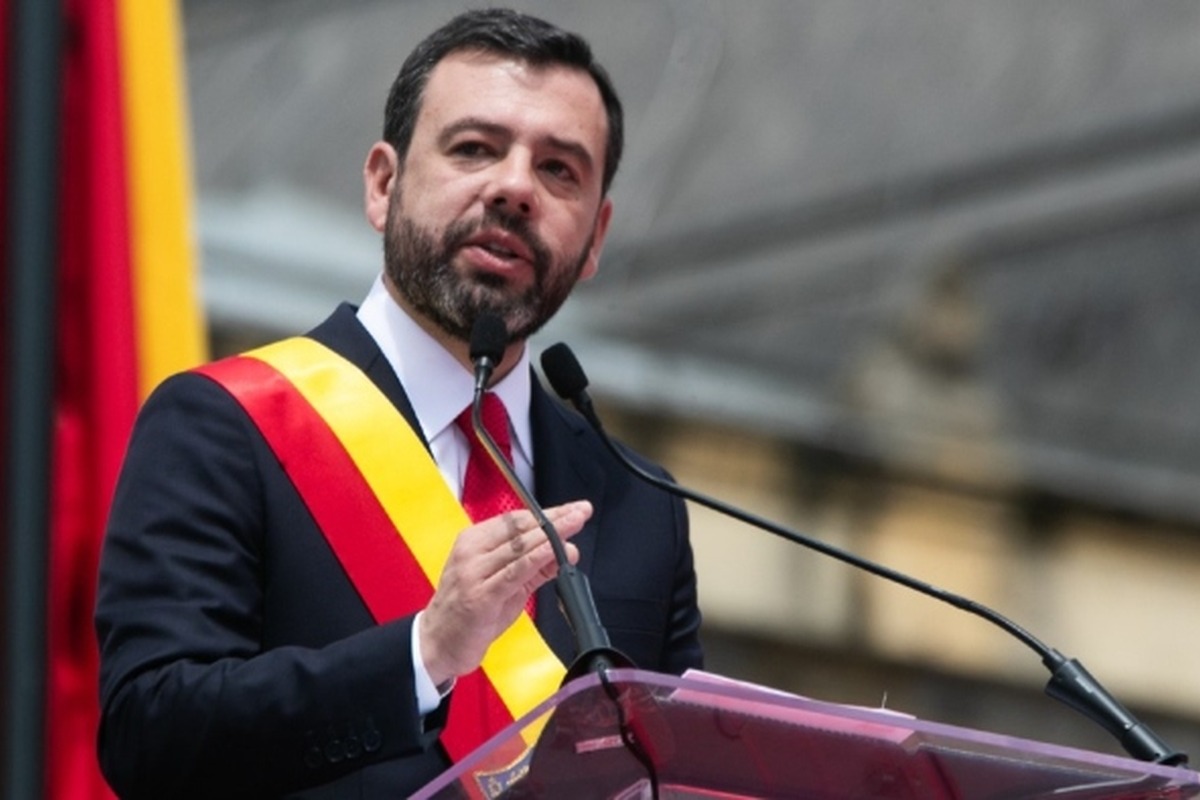Hombre de traje oscuro y corbata roja habla en un podio con micrófono, lleva banda diagonal roja y amarilla; al fondo se ve una bandera vertical con los mismos colores.