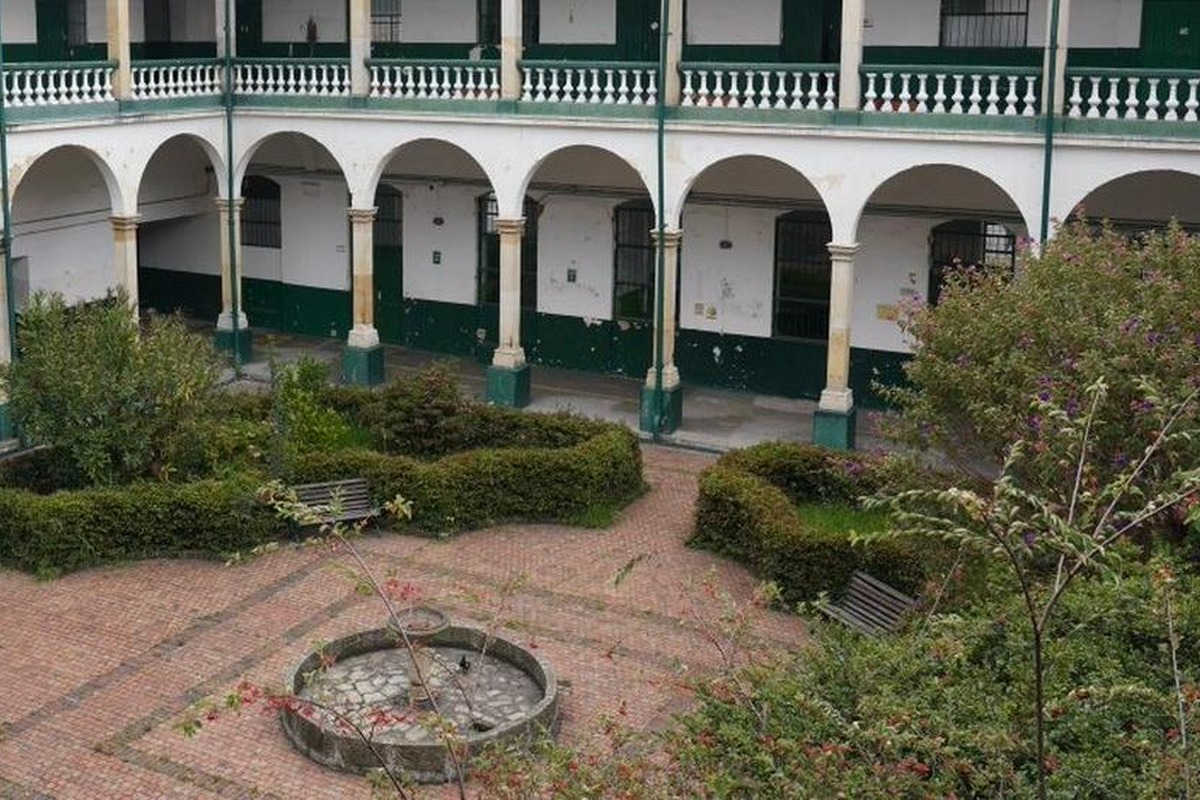 Patio interior con piso de ladrillo, fuente circular vacía al centro, bancas metálicas, setos y arbustos; alrededor, edificio de dos pisos con arcos y barandales blancos.