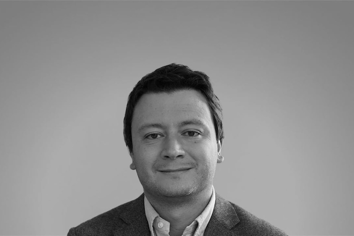 Retrato en blanco y negro de una persona mirando a la cámara, expresión neutra, cabello corto, chaqueta y camisa de cuello; fondo liso y claro sin otros objetos ni texto.