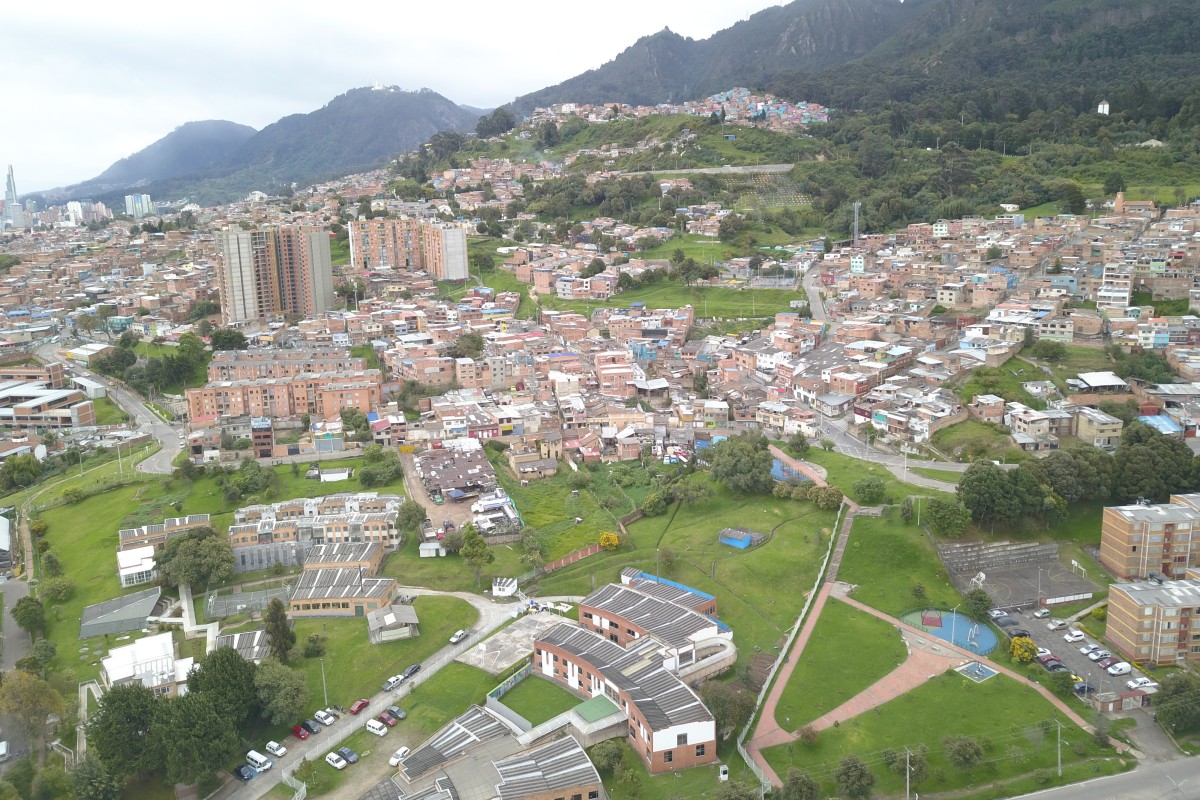 Vista aérea de un barrio con casas, edificios, calles, zonas verdes y montañas al fondo.