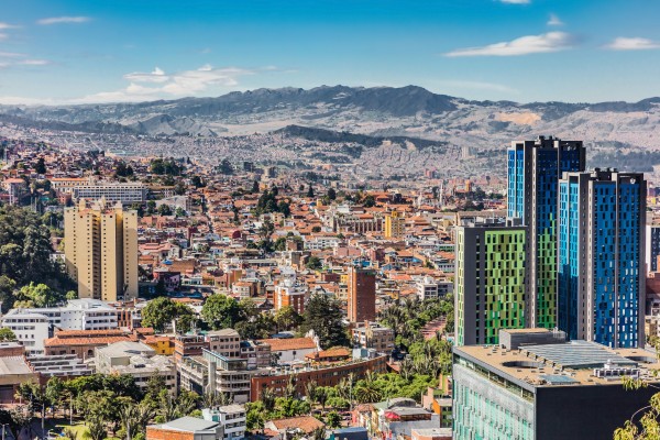 Vista panorámica de una ciudad con edificios, viviendas, árboles y montañas al fondo bajo un cielo azul.