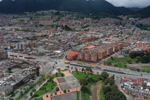 Vista aérea de una zona urbana con edificios, vías, parque con canchas deportivas, árboles, un canal y montañas al fondo.