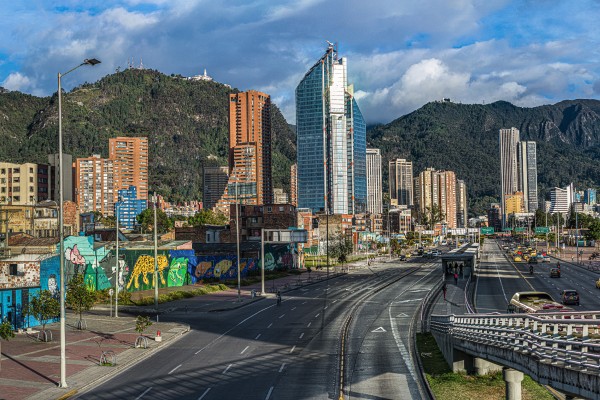 Vista urbana con una avenida amplia, edificios altos, grafitis, árboles y montañas al fondo bajo un cielo parcialmente nublado.
