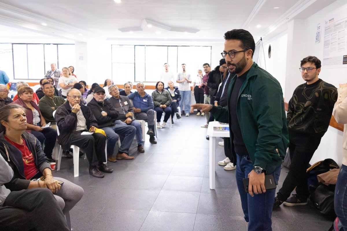 Hombre de pie gesticula ante un grupo de personas sentadas en una sala iluminada; varias personas observan.